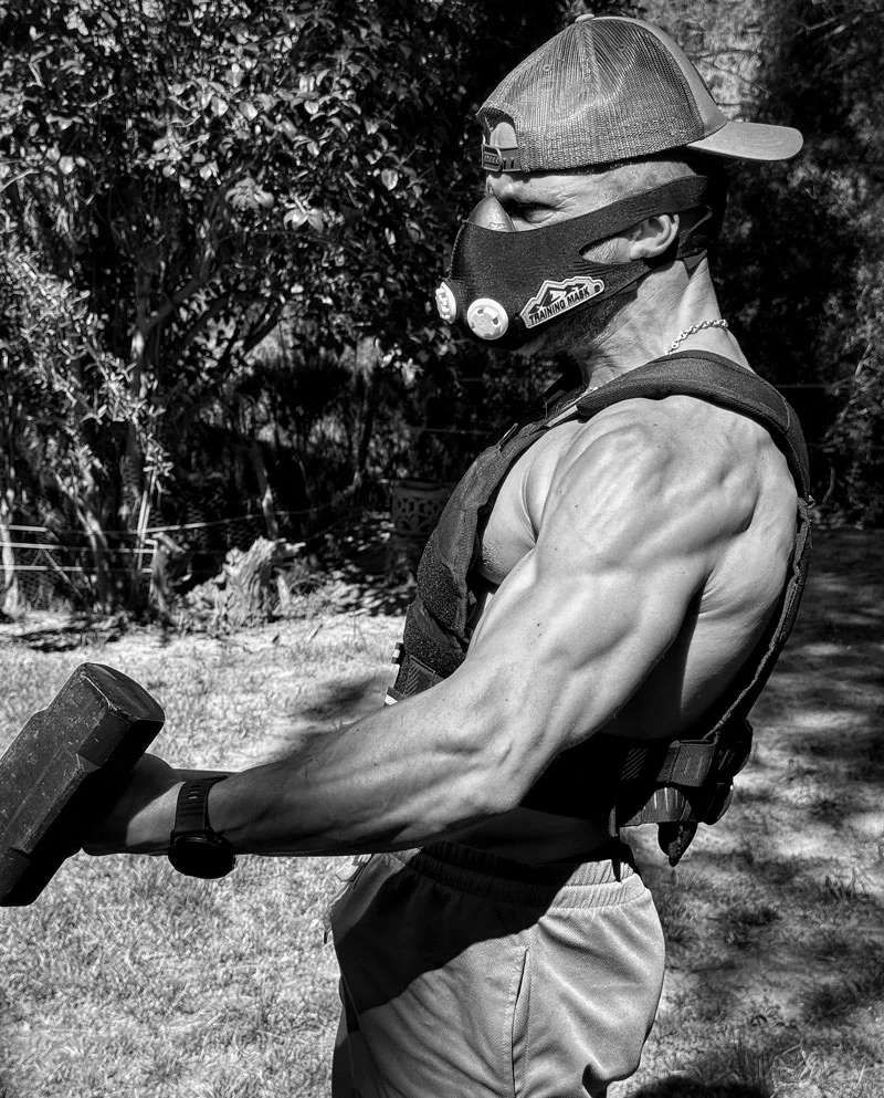 Hybrid athlete training outdoors with dumbbell, weighted vest and altitude mask, representing strength, endurance, and disciplined physical conditioning.