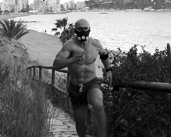 Hybrid athlete sprinting uphill by the sea wearing an altitude mask, symbolizing endurance, grit, and mental resilience through discomfort.