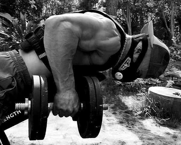 Athlete performing heavy dumbbell rows outdoors with training mask and weighted vest, showcasing raw strength, focus, and functional power.